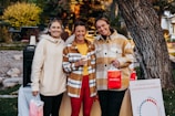 Three people are standing outdoors in a casual setting, smiling and holding shopping bags with the word 'MAREA' written on them. They are dressed in warm clothing, with two of them wearing plaid jackets. Behind them, there is greenery and trees, and a poster board with some text related to menstrual cycles.