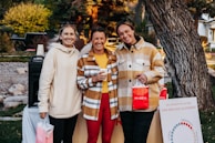 Three people are standing outdoors in a casual setting, smiling and holding shopping bags with the word 'MAREA' written on them. They are dressed in warm clothing, with two of them wearing plaid jackets. Behind them, there is greenery and trees, and a poster board with some text related to menstrual cycles.