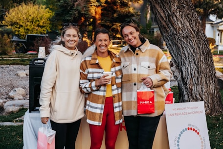 Three people are standing outdoors in a casual setting, smiling and holding shopping bags with the word 'MAREA' written on them. They are dressed in warm clothing, with two of them wearing plaid jackets. Behind them, there is greenery and trees, and a poster board with some text related to menstrual cycles.