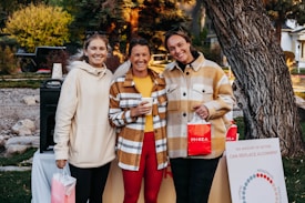 Three people are standing outdoors in a casual setting, smiling and holding shopping bags with the word 'MAREA' written on them. They are dressed in warm clothing, with two of them wearing plaid jackets. Behind them, there is greenery and trees, and a poster board with some text related to menstrual cycles.