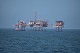 An offshore oil platform surrounded by the sea, featuring various structures such as cranes, pipes, and storage facilities. The platform stands on stilts above the water, with a clear blue sky in the background.