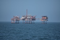 An offshore oil platform surrounded by the sea, featuring various structures such as cranes, pipes, and storage facilities. The platform stands on stilts above the water, with a clear blue sky in the background.