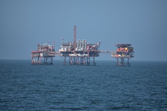 An offshore oil platform surrounded by the sea, featuring various structures such as cranes, pipes, and storage facilities. The platform stands on stilts above the water, with a clear blue sky in the background.