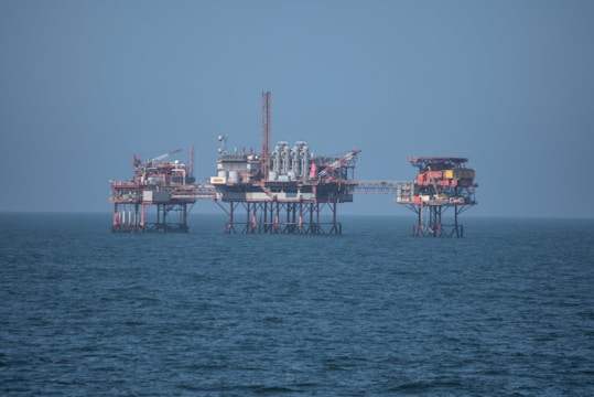 An offshore oil platform surrounded by the sea, featuring various structures such as cranes, pipes, and storage facilities. The platform stands on stilts above the water, with a clear blue sky in the background.