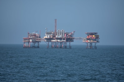 An offshore oil platform surrounded by the sea, featuring various structures such as cranes, pipes, and storage facilities. The platform stands on stilts above the water, with a clear blue sky in the background.