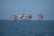 An offshore oil platform surrounded by the sea, featuring various structures such as cranes, pipes, and storage facilities. The platform stands on stilts above the water, with a clear blue sky in the background.
