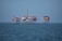 An offshore oil platform surrounded by the sea, featuring various structures such as cranes, pipes, and storage facilities. The platform stands on stilts above the water, with a clear blue sky in the background.