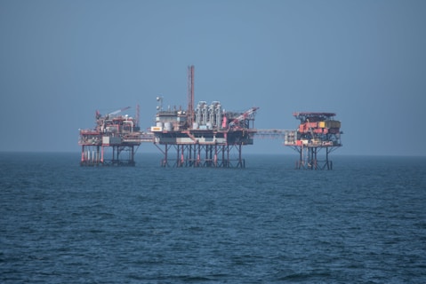 An offshore oil platform surrounded by the sea, featuring various structures such as cranes, pipes, and storage facilities. The platform stands on stilts above the water, with a clear blue sky in the background.