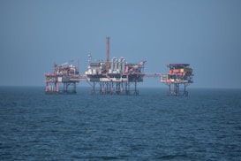 An offshore oil platform surrounded by the sea, featuring various structures such as cranes, pipes, and storage facilities. The platform stands on stilts above the water, with a clear blue sky in the background.
