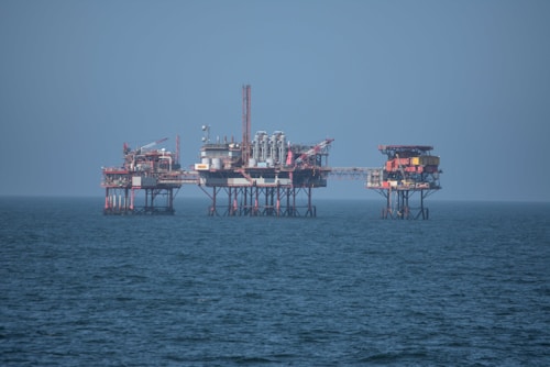An offshore oil platform surrounded by the sea, featuring various structures such as cranes, pipes, and storage facilities. The platform stands on stilts above the water, with a clear blue sky in the background.