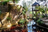 A serene natural setting with a rustic wooden structure featuring a thatched roof perched above a rocky landscape. Lush greenery, including various plants and palm trees, adorns the area. A small pond or stream with clear water reflects the surrounding vegetation and structures. Sunlight filters through the trees, casting a gentle light on the scene.