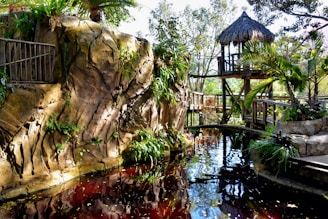 A serene natural setting with a rustic wooden structure featuring a thatched roof perched above a rocky landscape. Lush greenery, including various plants and palm trees, adorns the area. A small pond or stream with clear water reflects the surrounding vegetation and structures. Sunlight filters through the trees, casting a gentle light on the scene.