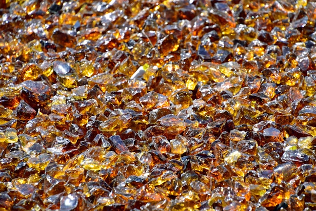 A large collection of amber-colored glass shards scattered closely together. The pieces have various sizes and shapes, reflecting light and creating a shimmering effect. There are hints of darker and lighter shades within the glass, enhancing the sparkling appearance.