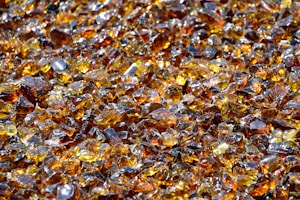 A large collection of amber-colored glass shards scattered closely together. The pieces have various sizes and shapes, reflecting light and creating a shimmering effect. There are hints of darker and lighter shades within the glass, enhancing the sparkling appearance.
