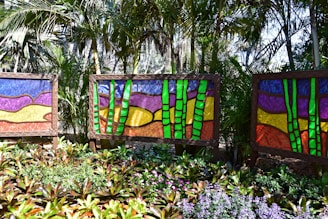Vibrant stained glass art panels are displayed outdoors amidst dense tropical foliage. Each panel features abstract designs with vivid colors, primarily green bamboo stalks juxtaposed against backgrounds of purple, yellow, and red. The foreground is filled with lush green plants and assorted flowers adding to the lively ambiance of the scene.