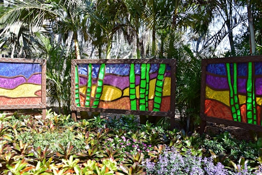 Vibrant stained glass art panels are displayed outdoors amidst dense tropical foliage. Each panel features abstract designs with vivid colors, primarily green bamboo stalks juxtaposed against backgrounds of purple, yellow, and red. The foreground is filled with lush green plants and assorted flowers adding to the lively ambiance of the scene.