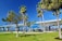 A scenic park area featuring lush green grass and tall palm trees under a clear blue sky. A large bridge spans the background, with a pavilion with a bright blue roof in the foreground. There are people visible under the pavilion and nearby, engaged in leisure activities.