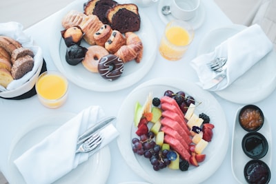 A beautifully arranged breakfast spread featuring a variety of pastries, including croissants and donuts, and a vibrant fruit platter with watermelon, grapes, and berries. Accompanying the meal are glasses of orange juice and small bowls of assorted jams. Tableware includes plates, utensils, and napkins, creating an elegant dining setting.