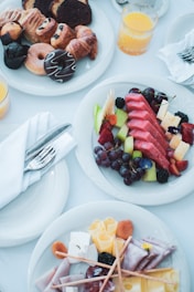A selection of breakfast items is arranged on a table, including a plate with varied pastries such as donuts, croissants, and slices of cake. Another plate is filled with colorful fruits like watermelon, grapes, and pieces of cheese. A plate with assorted meats, cheeses, and breadsticks is also present. Glasses of orange juice and neatly placed cutlery wrapped in napkins complement the scene, creating an inviting and elegant breakfast setting.