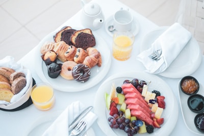 A breakfast spread featuring a variety of pastries like muffins, croissants, and donuts on a white plate. Next to it, a colorful fruit platter with slices of watermelon, pineapple, grapes, and strawberries. Accompanying these are glasses of orange juice, a teapot, and a small basket of bread. There are also small dishes with jams and a neatly placed napkin with cutlery.