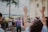 A group of diverse people is gathered outdoors, some with their hands raised, creating a lively and engaging atmosphere. The setting features a tall palm tree and part of a stone building, suggesting a warm, tropical location. The mood appears to be joyful and celebratory.