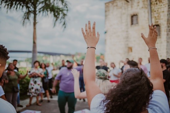 A group of diverse people is gathered outdoors, some with their hands raised, creating a lively and engaging atmosphere. The setting features a tall palm tree and part of a stone building, suggesting a warm, tropical location. The mood appears to be joyful and celebratory.