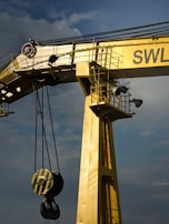 A large industrial crane painted in yellow dominates the image. It features a sturdy, vertical beam with an attached pulley system that includes a heavy-duty hook. The crane is positioned against a cloudy sky, adding to the industrial feel. The letters 'SWL' are partially visible on the crane.