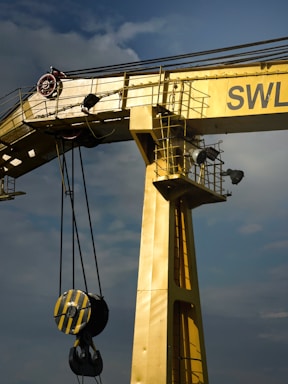 A large industrial crane painted in yellow dominates the image. It features a sturdy, vertical beam with an attached pulley system that includes a heavy-duty hook. The crane is positioned against a cloudy sky, adding to the industrial feel. The letters 'SWL' are partially visible on the crane.