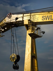 A large industrial crane painted in yellow dominates the image. It features a sturdy, vertical beam with an attached pulley system that includes a heavy-duty hook. The crane is positioned against a cloudy sky, adding to the industrial feel. The letters 'SWL' are partially visible on the crane.
