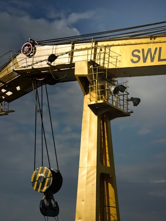 A large industrial crane painted in yellow dominates the image. It features a sturdy, vertical beam with an attached pulley system that includes a heavy-duty hook. The crane is positioned against a cloudy sky, adding to the industrial feel. The letters 'SWL' are partially visible on the crane.