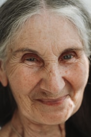 An elderly person with gray hair gazes forward with a gentle expression. The face is close-up, showing fine wrinkles and a soft smile, conveying a sense of wisdom and warmth.