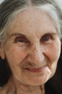 An elderly person with gray hair gazes forward with a gentle expression. The face is close-up, showing fine wrinkles and a soft smile, conveying a sense of wisdom and warmth.