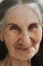 An elderly person with gray hair gazes forward with a gentle expression. The face is close-up, showing fine wrinkles and a soft smile, conveying a sense of wisdom and warmth.