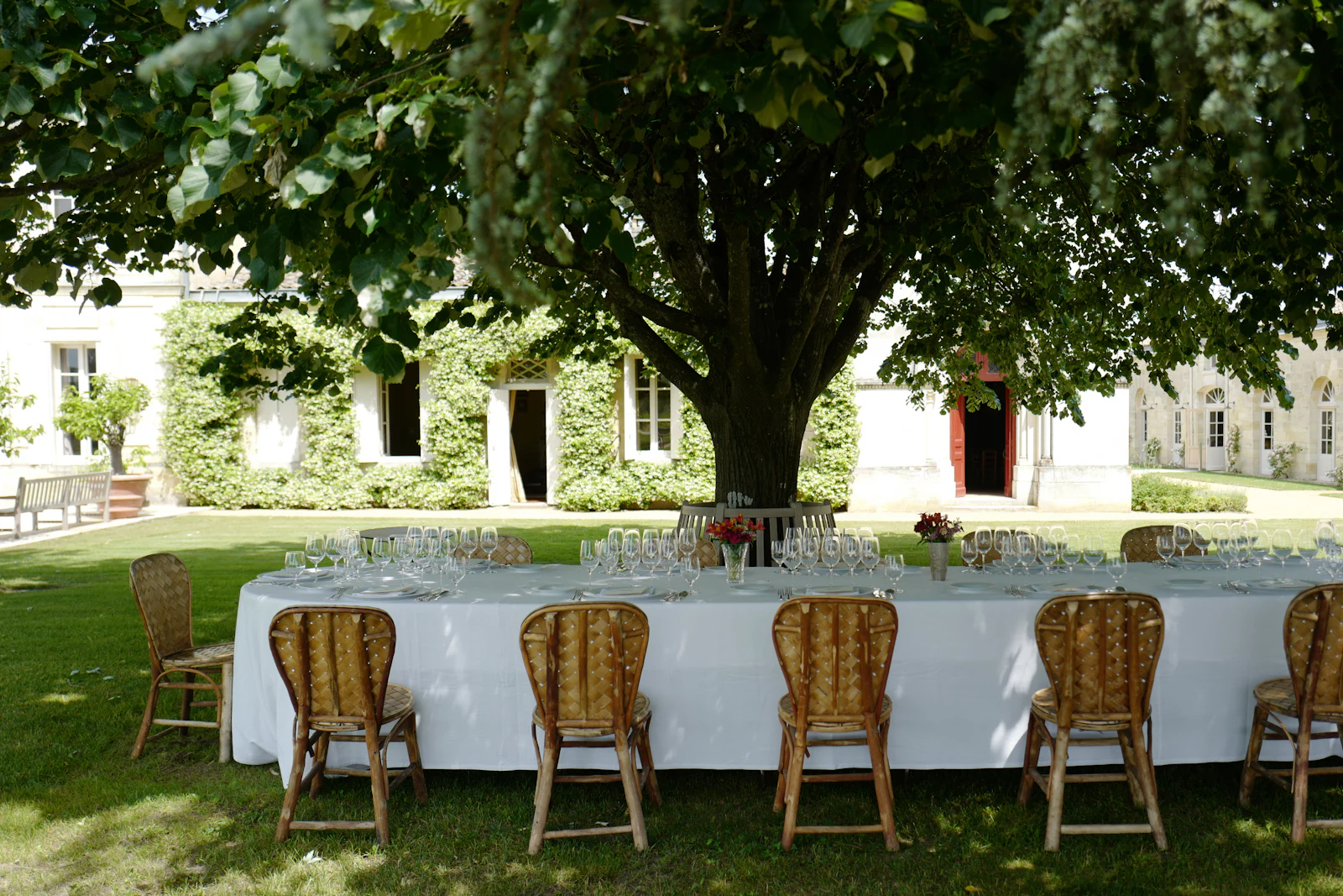 A long table in white linen laid under a large tree, in front of a stone house.