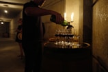 A person pours wine from a green bottle into multiple wine glasses arranged on a wooden barrel. The setting appears to be dimly lit, possibly in a wine cellar, with other people in the background.
