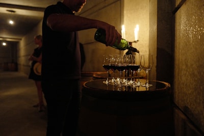 A person pours wine from a green bottle into multiple wine glasses arranged on a wooden barrel. The setting appears to be dimly lit, possibly in a wine cellar, with other people in the background.