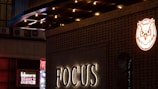 A night scene featuring a building facade illuminated with a sign that reads 'FOCUS'. Above it, there's a glowing owl logo in orange. To the left, additional signage in various colors, including red and white, adds a vibrant touch. The architectural design includes a gridded pattern with lighting that accentuates the structure.
