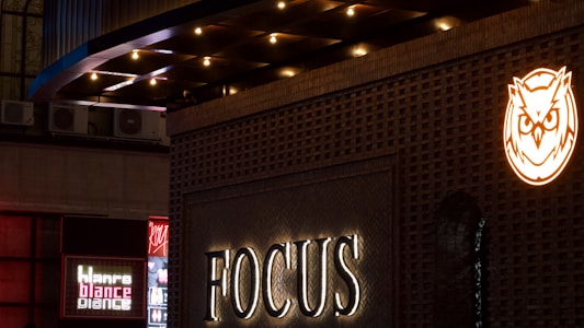 A night scene featuring a building facade illuminated with a sign that reads 'FOCUS'. Above it, there's a glowing owl logo in orange. To the left, additional signage in various colors, including red and white, adds a vibrant touch. The architectural design includes a gridded pattern with lighting that accentuates the structure.