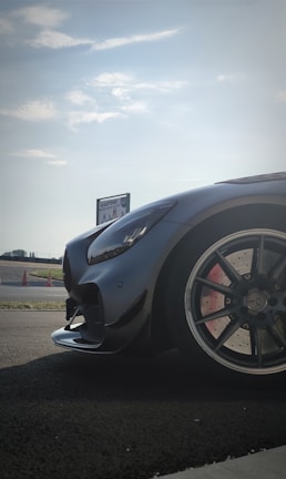 A sleek, high-performance car is parked on a racetrack. The focus is on the car's front, showcasing its glossy black exterior and stylish design. The background features a clear sky and a sign that reads 'KARTING.' Traffic cones and asphalt further indicate a racing environment.