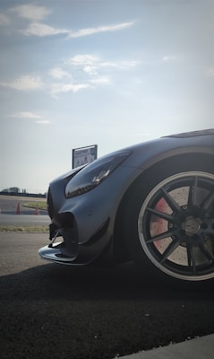 A sleek, high-performance car is parked on a racetrack. The focus is on the car's front, showcasing its glossy black exterior and stylish design. The background features a clear sky and a sign that reads 'KARTING.' Traffic cones and asphalt further indicate a racing environment.