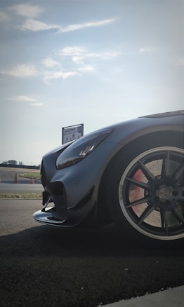 A sleek, high-performance car is parked on a racetrack. The focus is on the car's front, showcasing its glossy black exterior and stylish design. The background features a clear sky and a sign that reads 'KARTING.' Traffic cones and asphalt further indicate a racing environment.