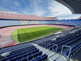 A vast football stadium with empty blue and red seats surrounding a well-maintained green pitch. The words 'MES QUE UN CLUB' are visible in yellow on the seating area. The sky is partly cloudy with a curved roof partially covering the stands.