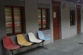 Four colorful plastic chairs are lined up outside a building with concrete walls. The wall features two windows on the left with brown frames and a wooden door on the right. Light gray tiles are present on the ground, and a red train is faintly visible through the window reflection.