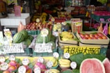 A vibrant fruit market stall displaying a variety of fresh produce, including watermelons, bananas, apples, dragon fruits, and other colorful fruits. Each type of fruit is organized in crates with price tags and signs in multiple languages, likely catering to customers in the area.