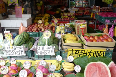 A vibrant fruit market stall displaying a variety of fresh produce, including watermelons, bananas, apples, dragon fruits, and other colorful fruits. Each type of fruit is organized in crates with price tags and signs in multiple languages, likely catering to customers in the area.