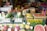 A vibrant fruit market stall displaying a variety of fresh produce, including watermelons, bananas, apples, dragon fruits, and other colorful fruits. Each type of fruit is organized in crates with price tags and signs in multiple languages, likely catering to customers in the area.