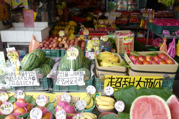 A vibrant fruit market stall displaying a variety of fresh produce, including watermelons, bananas, apples, dragon fruits, and other colorful fruits. Each type of fruit is organized in crates with price tags and signs in multiple languages, likely catering to customers in the area.