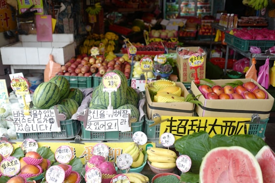 A vibrant fruit market stall displaying a variety of fresh produce, including watermelons, bananas, apples, dragon fruits, and other colorful fruits. Each type of fruit is organized in crates with price tags and signs in multiple languages, likely catering to customers in the area.