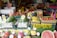 A vibrant fruit market stall displaying a variety of fresh produce, including watermelons, bananas, apples, dragon fruits, and other colorful fruits. Each type of fruit is organized in crates with price tags and signs in multiple languages, likely catering to customers in the area.