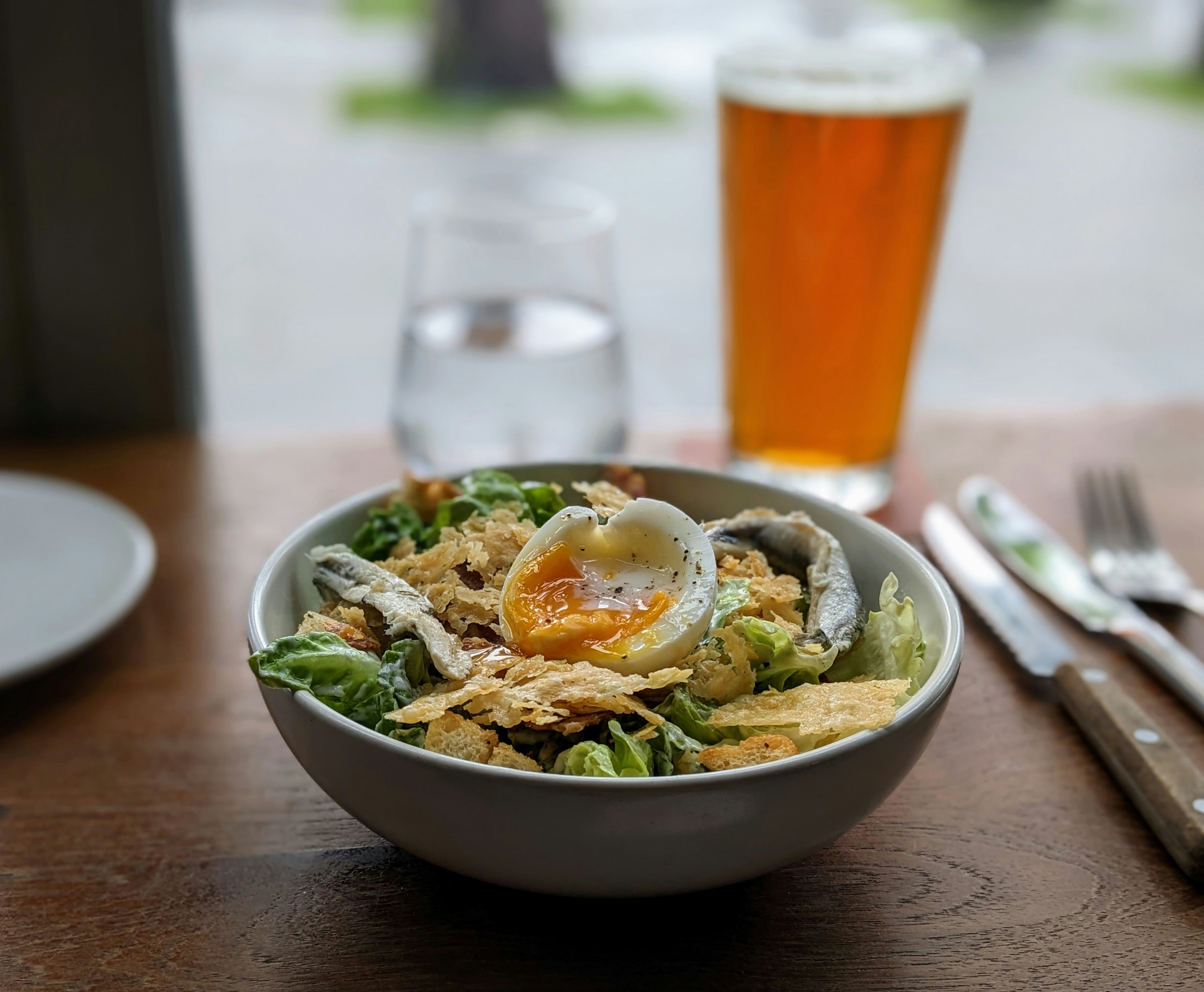 a bowl of food and a glass of beer on a table, Salad with egg and a beer in the background.